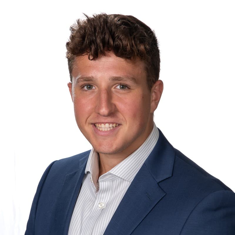 Smiling young man with wavy brown hair wearing a blue blazer and a patterned shirt.